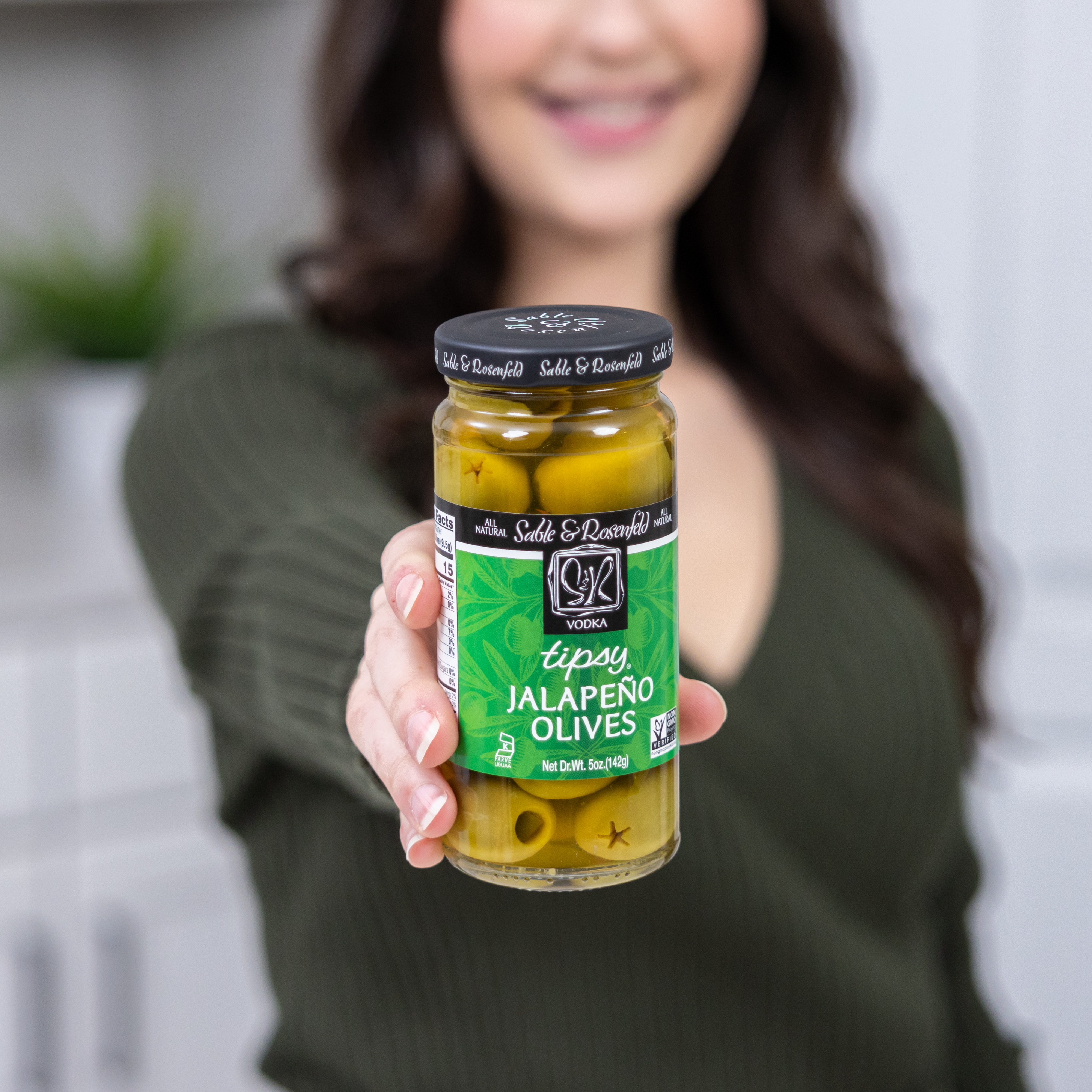 A woman holding a jar of Sable & Rosenfeld Tipsy Jalapeño Olives, featuring green olives stuffed with jalapeños, against a blurred kitchen background.