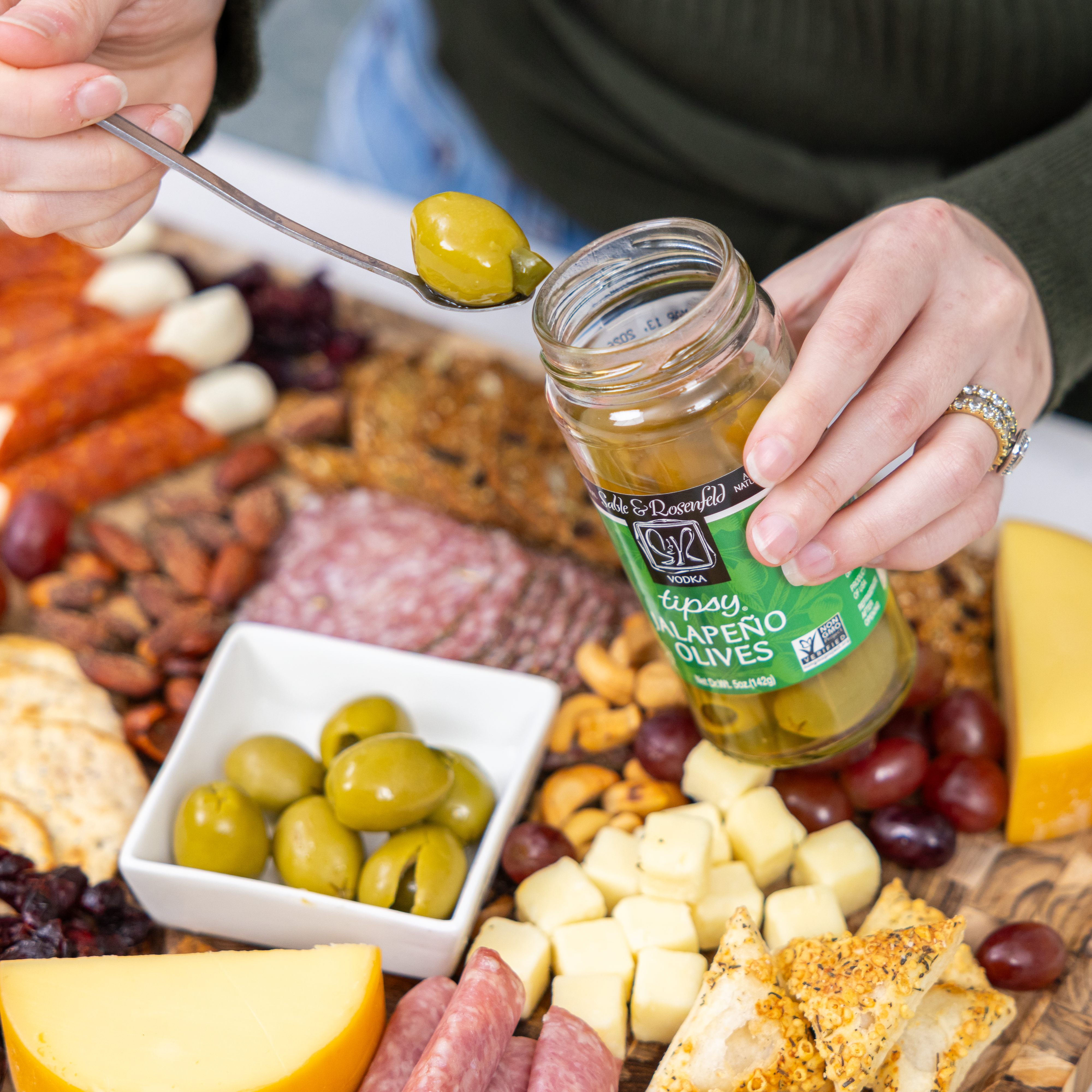 A person holding a jar of Sable & Rosenfeld Tipsy Jalapeño Olives while scooping out an olive with a spoon over a charcuterie board filled with cheese, nuts, grapes, and cured meats.