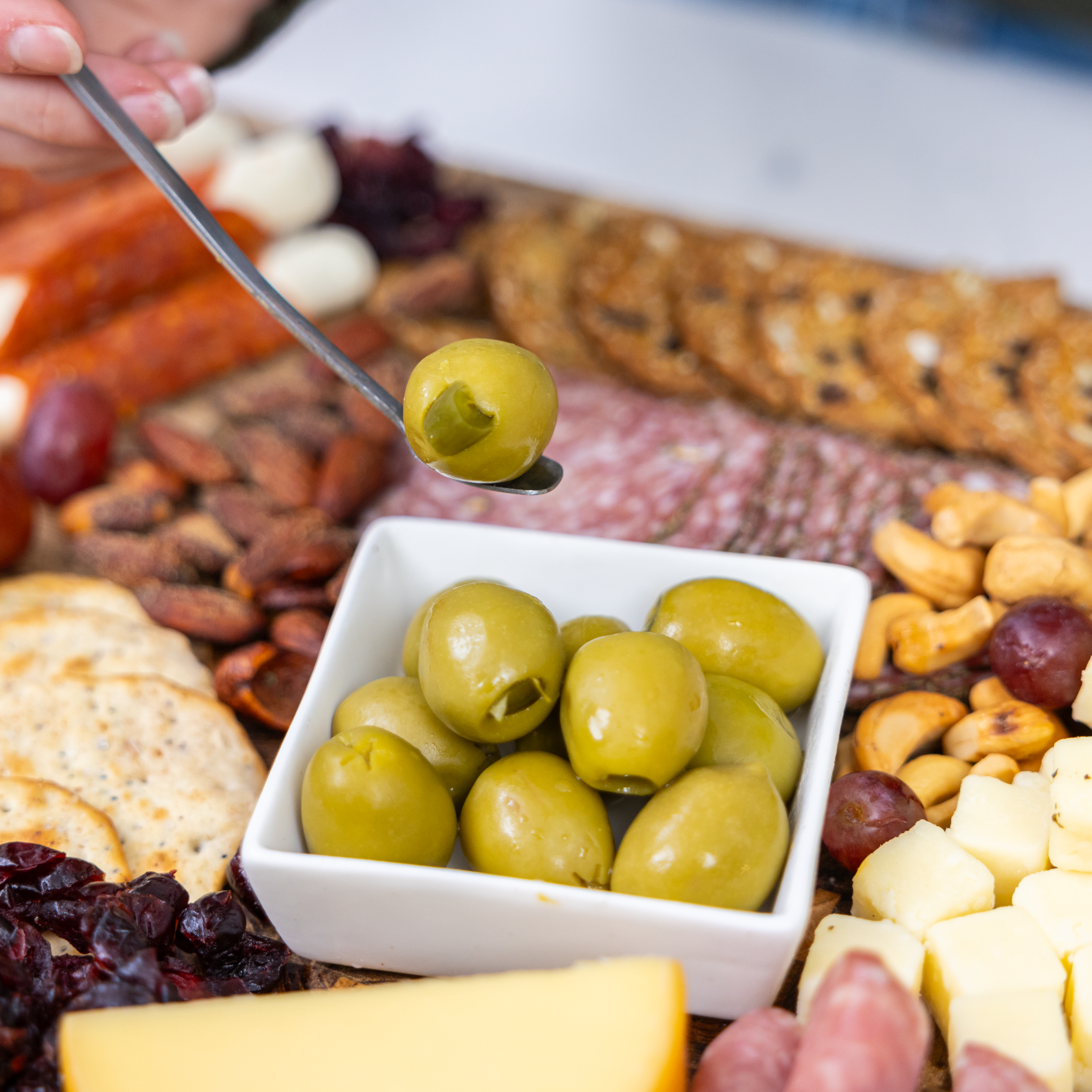 A close-up of a white bowl filled with jalapeño stuffed olives on a charcuterie board with cheese, nuts, grapes, crackers, and cured meats. A hand lifts an olive with a spoon.