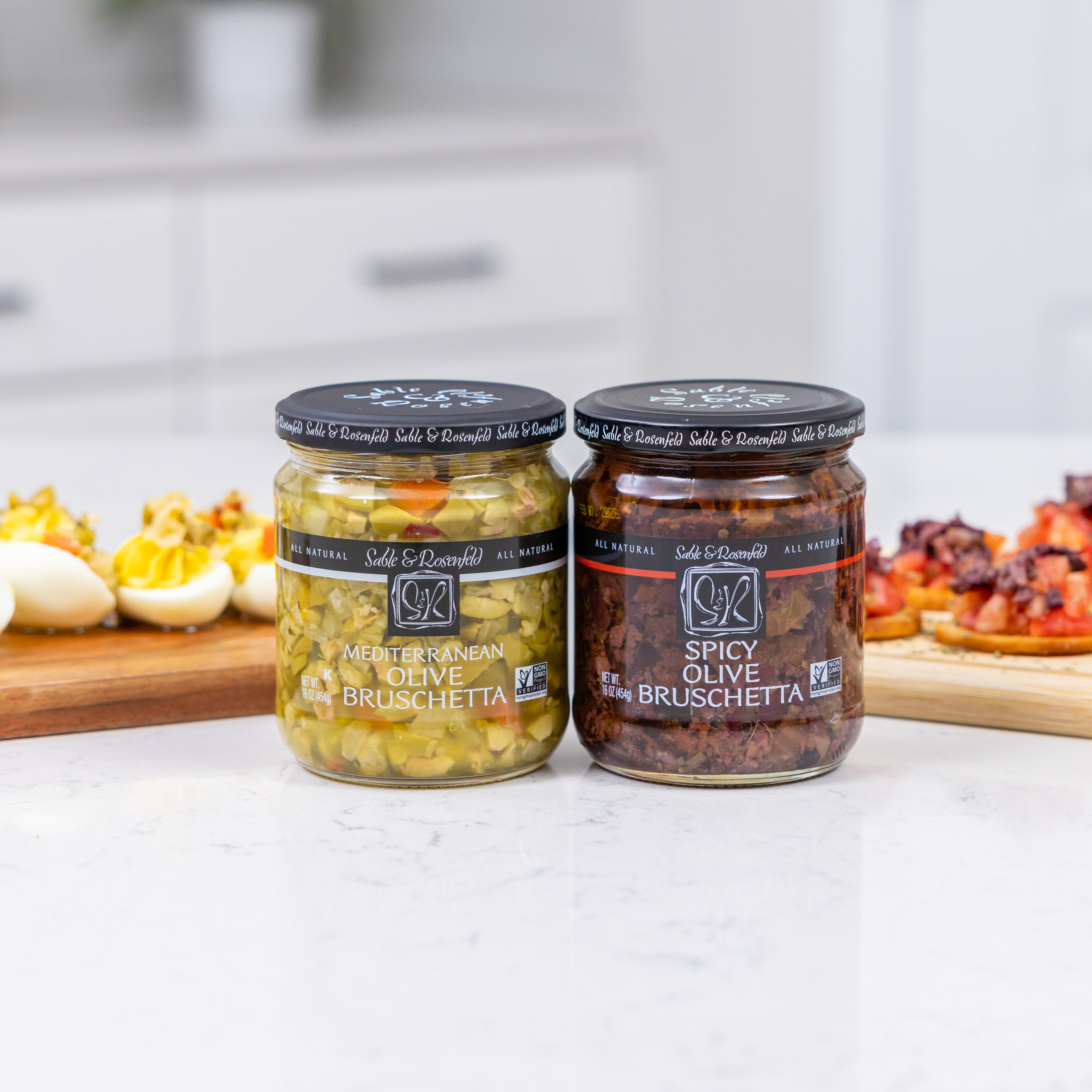 Two jars of Olive Bruschetta – Mediterranean Olive Bruschetta and Spicy Olive Bruschetta – placed on a white kitchen counter. In the background, deviled eggs and crispy bruschetta topped with diced tomatoes and bruschetta spread are displayed on wooden boards.