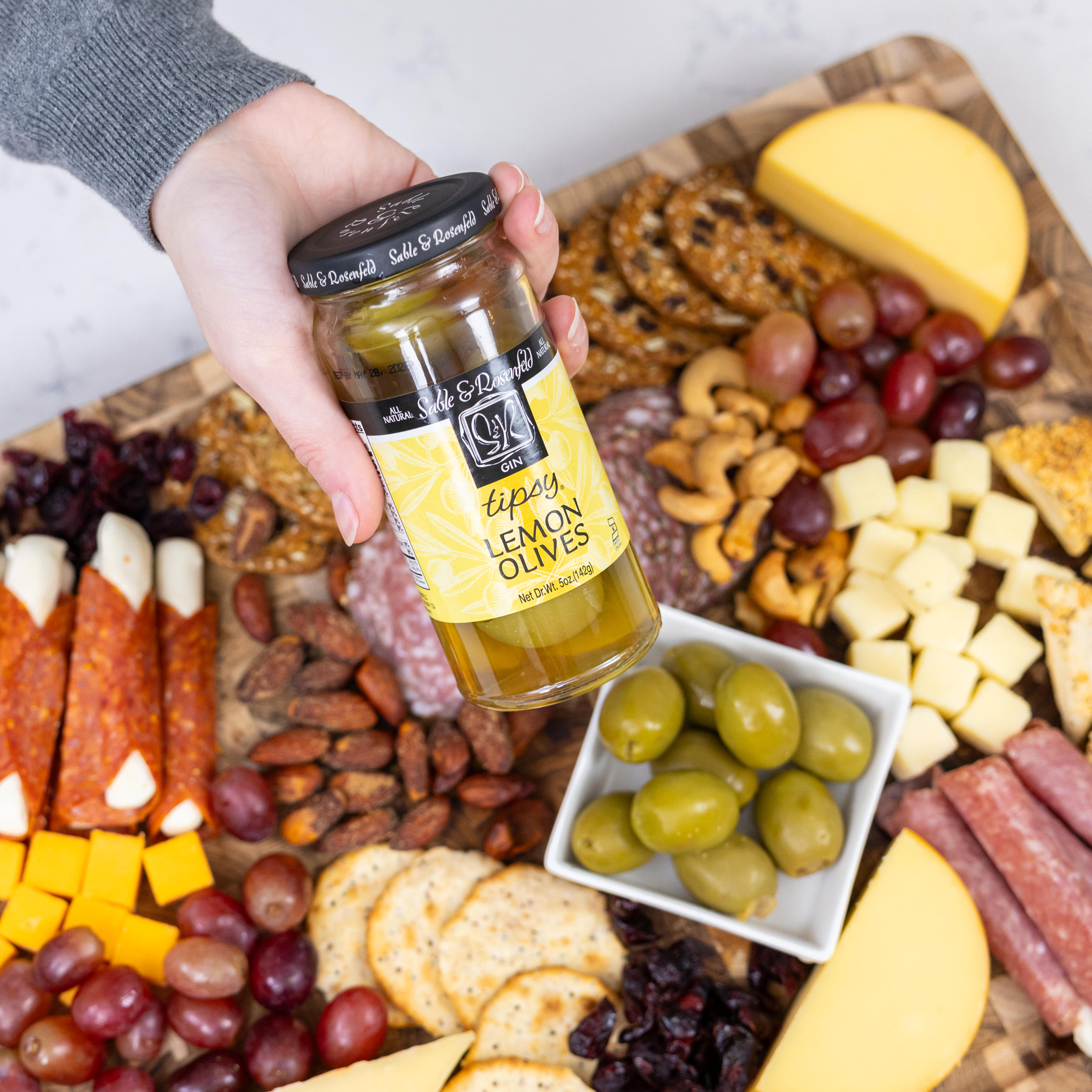 A hand holding a jar of Sable & Rosenfeld Tipsy Lemon Olives over a gourmet charcuterie board, featuring cheese, crackers, cured meats, grapes, nuts, and a dish of lemon-stuffed green olives.