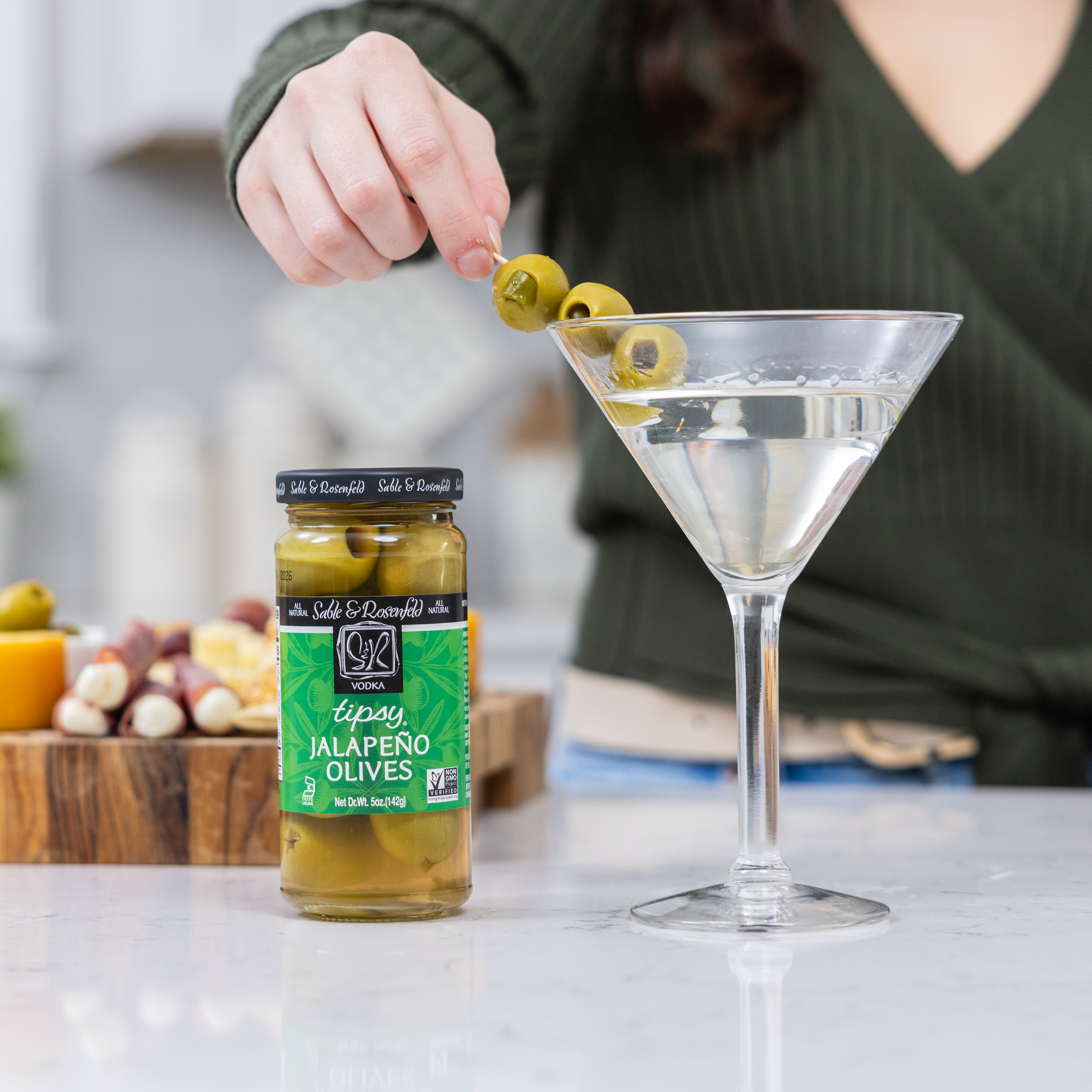 A person garnishing a martini with olives in vodka, using Sable & Rosenfeld Tipsy Jalapeño Olives. A jar of the olives is placed on a white countertop with a charcuterie board in the background.