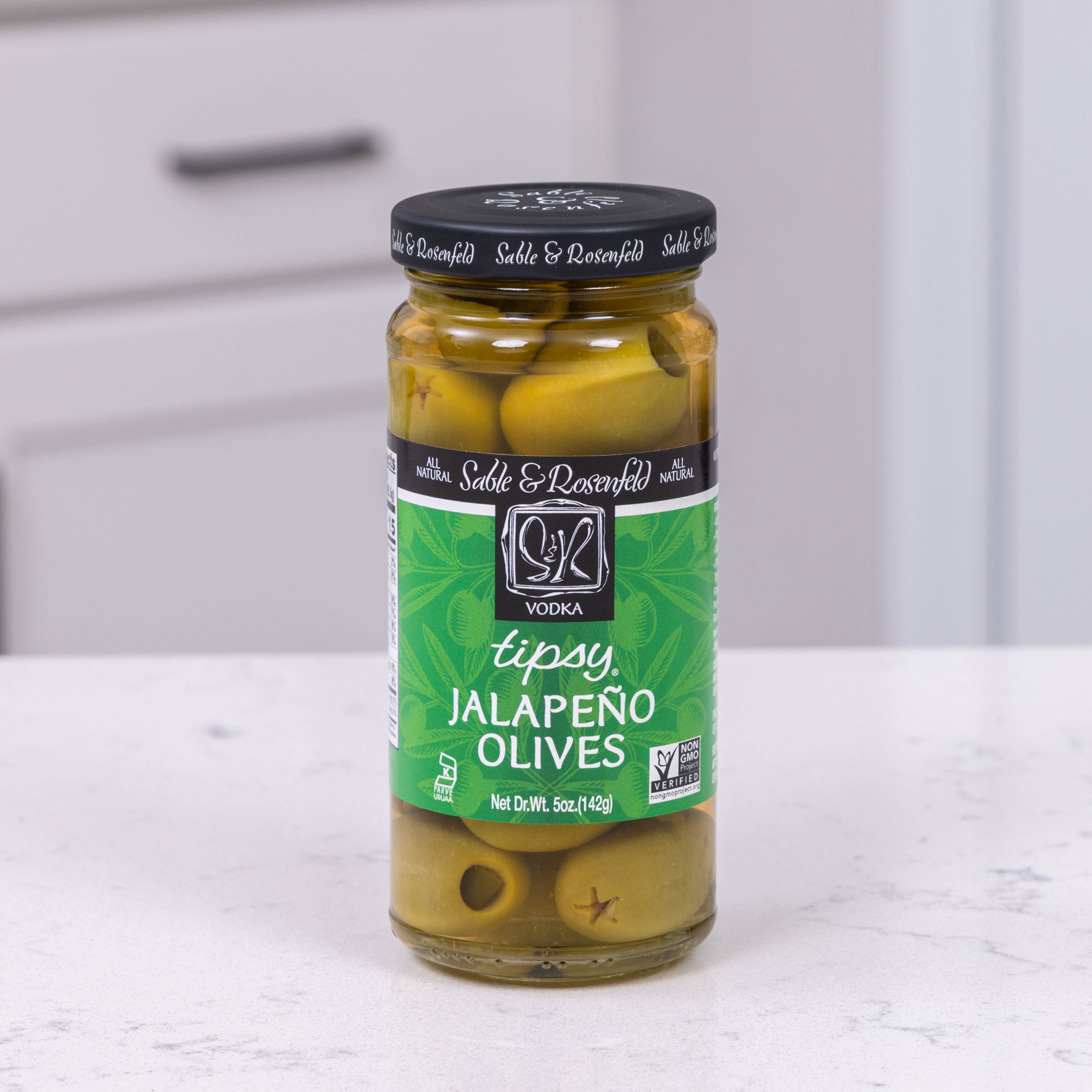 A jar of Sable & Rosenfeld Tipsy Olives with Jalapeño, featuring green olives stuffed with jalapeño peppers, placed on a white kitchen countertop.