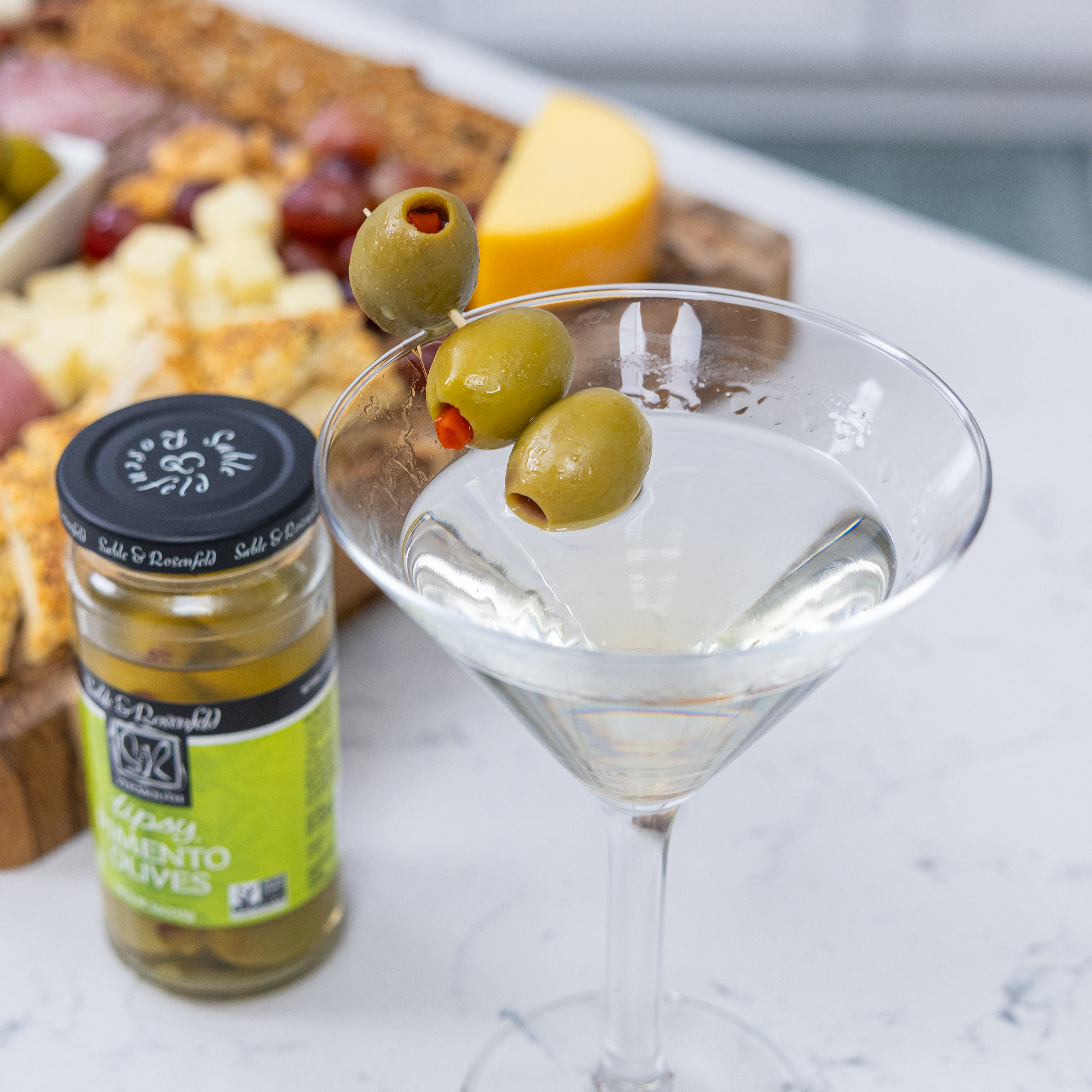 A martini garnished with three pimento-stuffed olives on a skewer, next to a jar of Sable & Rosenfeld Tipsy Pimento Olives and a charcuterie board with cheese, grapes, and crackers.