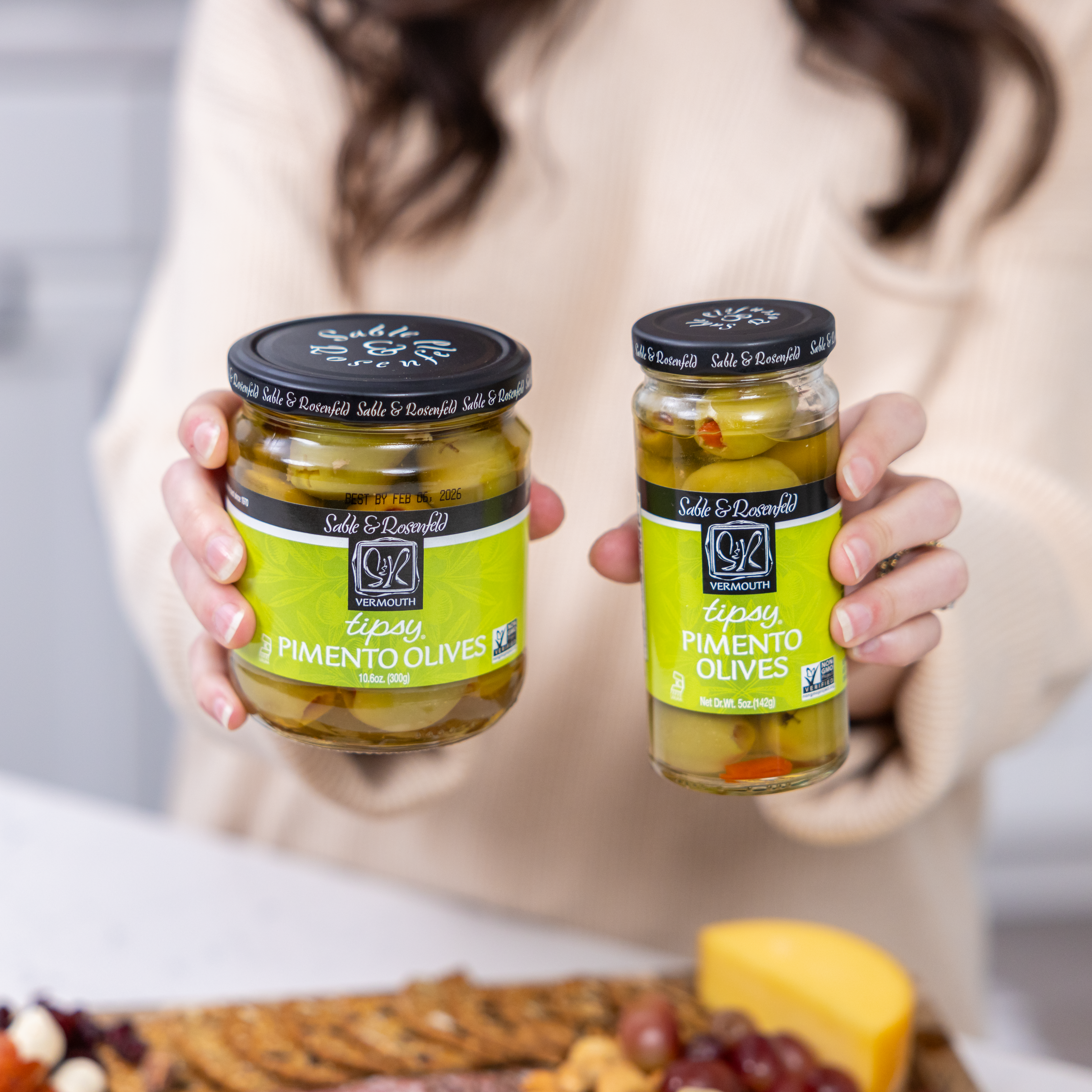 A woman holding two jars of Sable & Rosenfeld Tipsy Pimento Olives, featuring green olives stuffed with pimentos and infused with vermouth, in a bright kitchen setting.