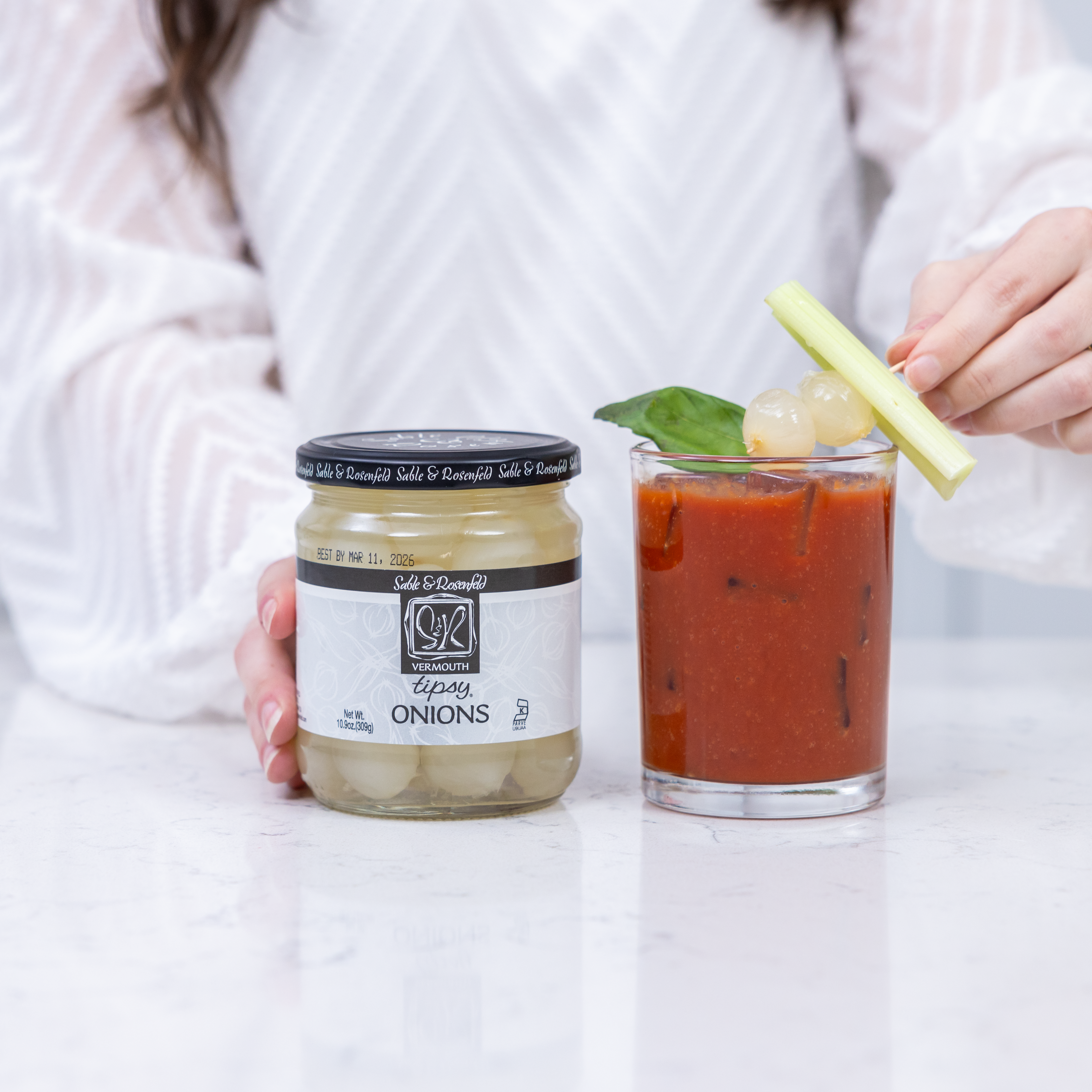 A jar of Sable & Rosenfeld Vermouth Tipsy Onions alongside a Bloody Mary cocktail garnished with silverskin onions on a celery stick. A person in a white sweater is placing the garnish on the drink.