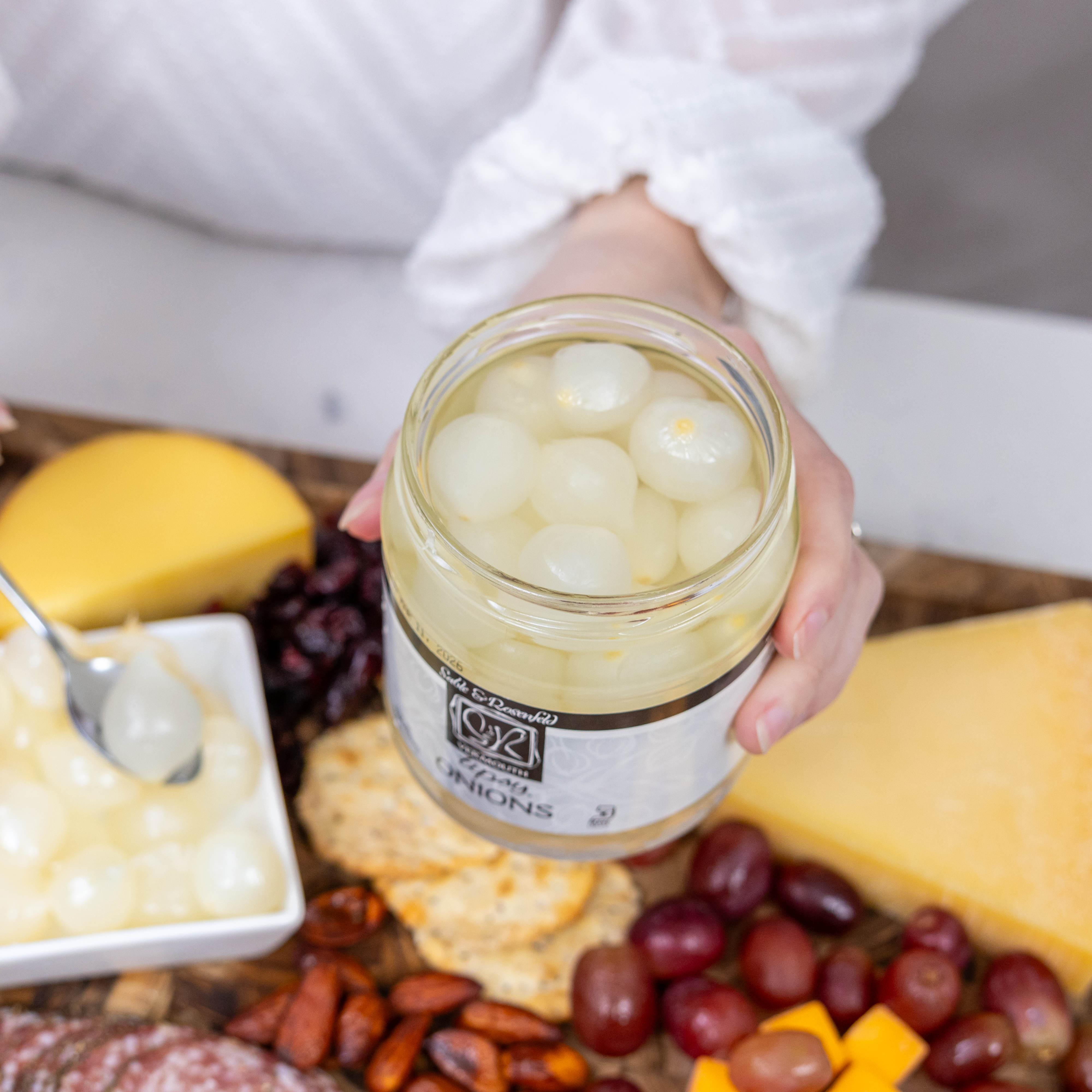 A close-up of an open jar of silverskin pickled onions soaked in vermouth, held by a person wearing a white sweater. The jar is placed above a charcuterie board with cheese, crackers, nuts, grapes, and a dish of pickled onions.