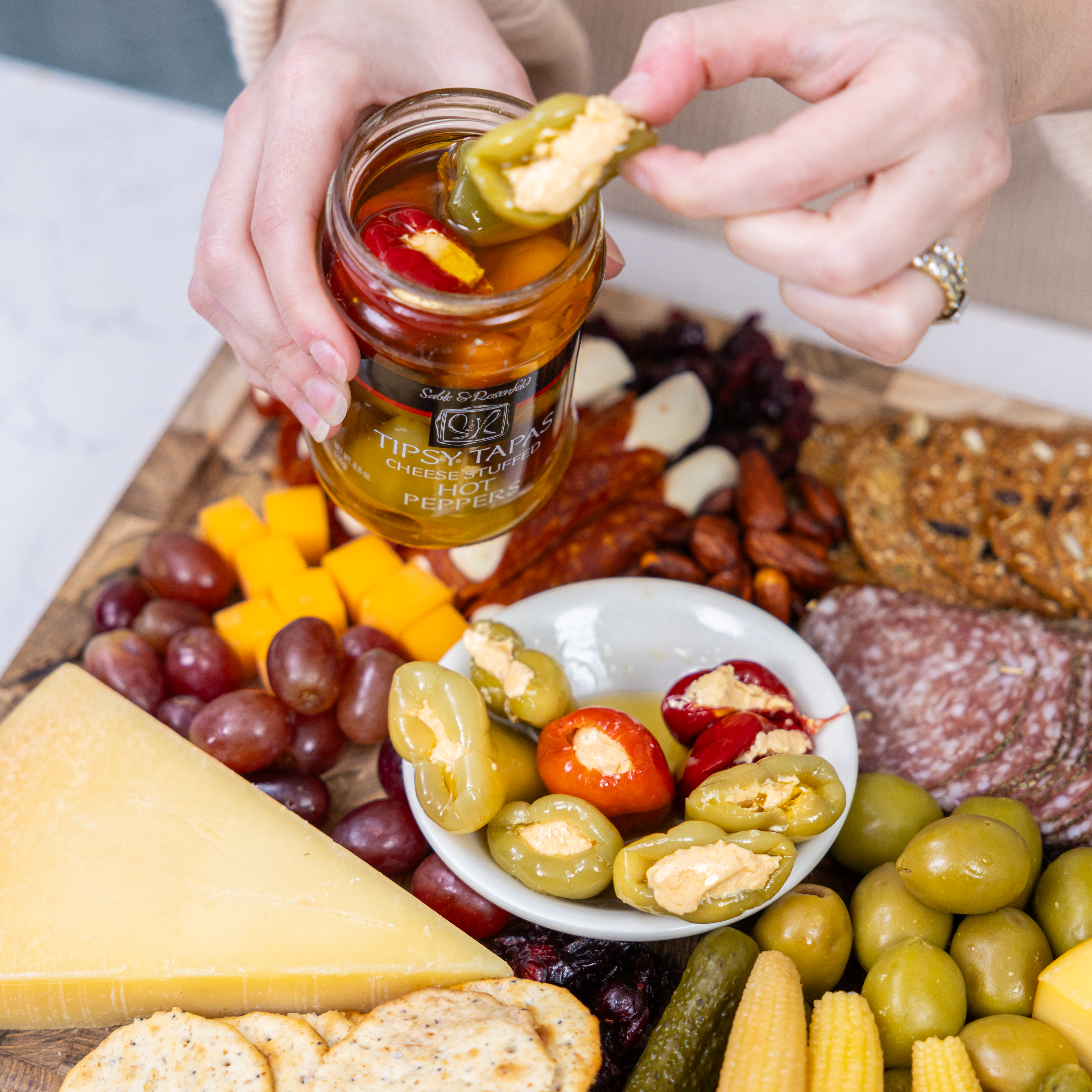 A person holding a jar of Sable & Rosenfeld Tipsy Tapas stuffed hot peppers with cream cheese, with a charcuterie board featuring cheese, olives, crackers, and cured meats in the background.