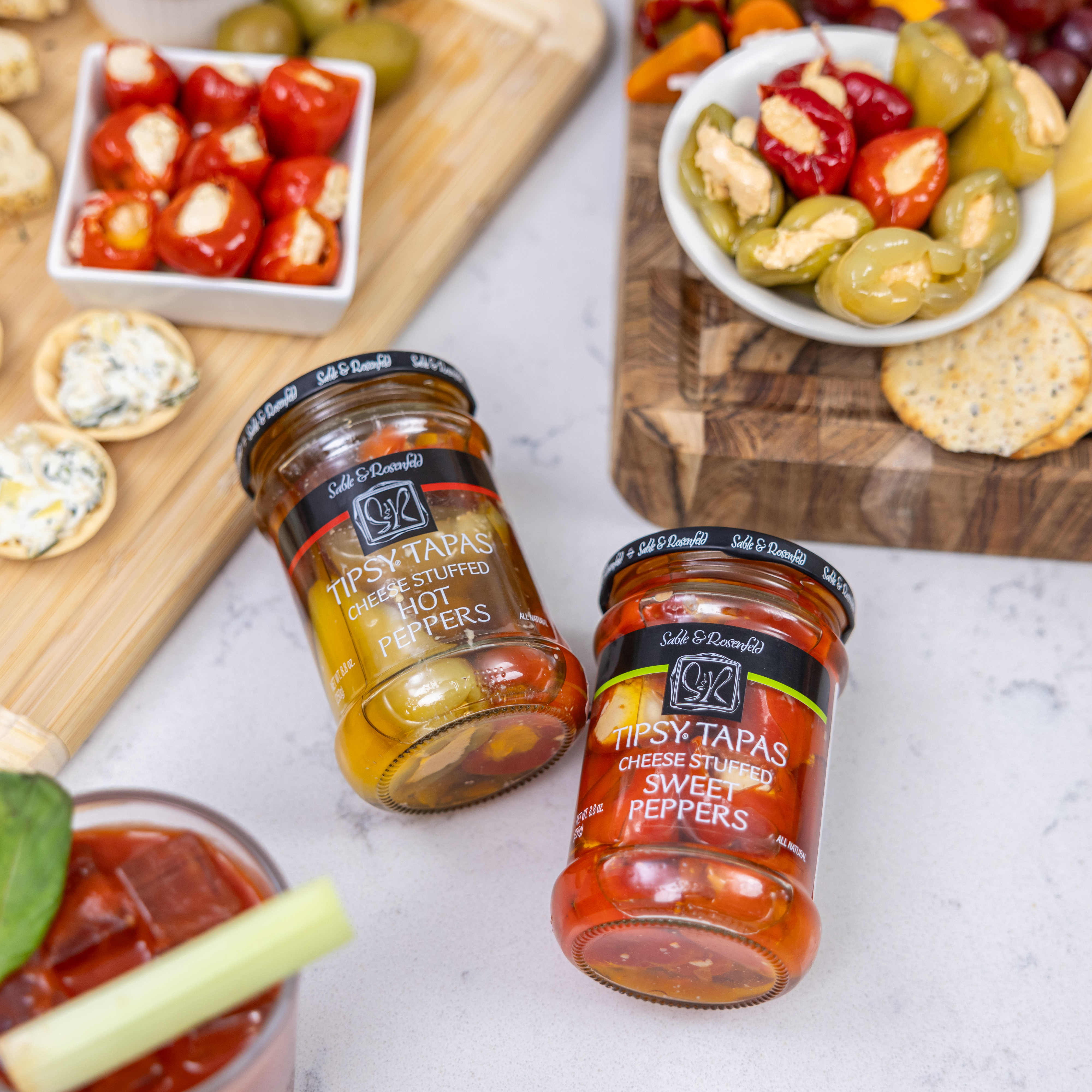 Jars of Sable & Rosenfeld Tipsy Tapas stuffed sweet peppers with cream cheese and hot peppers, displayed alongside a charcuterie board with cheese, crackers, and a Bloody Mary cocktail.