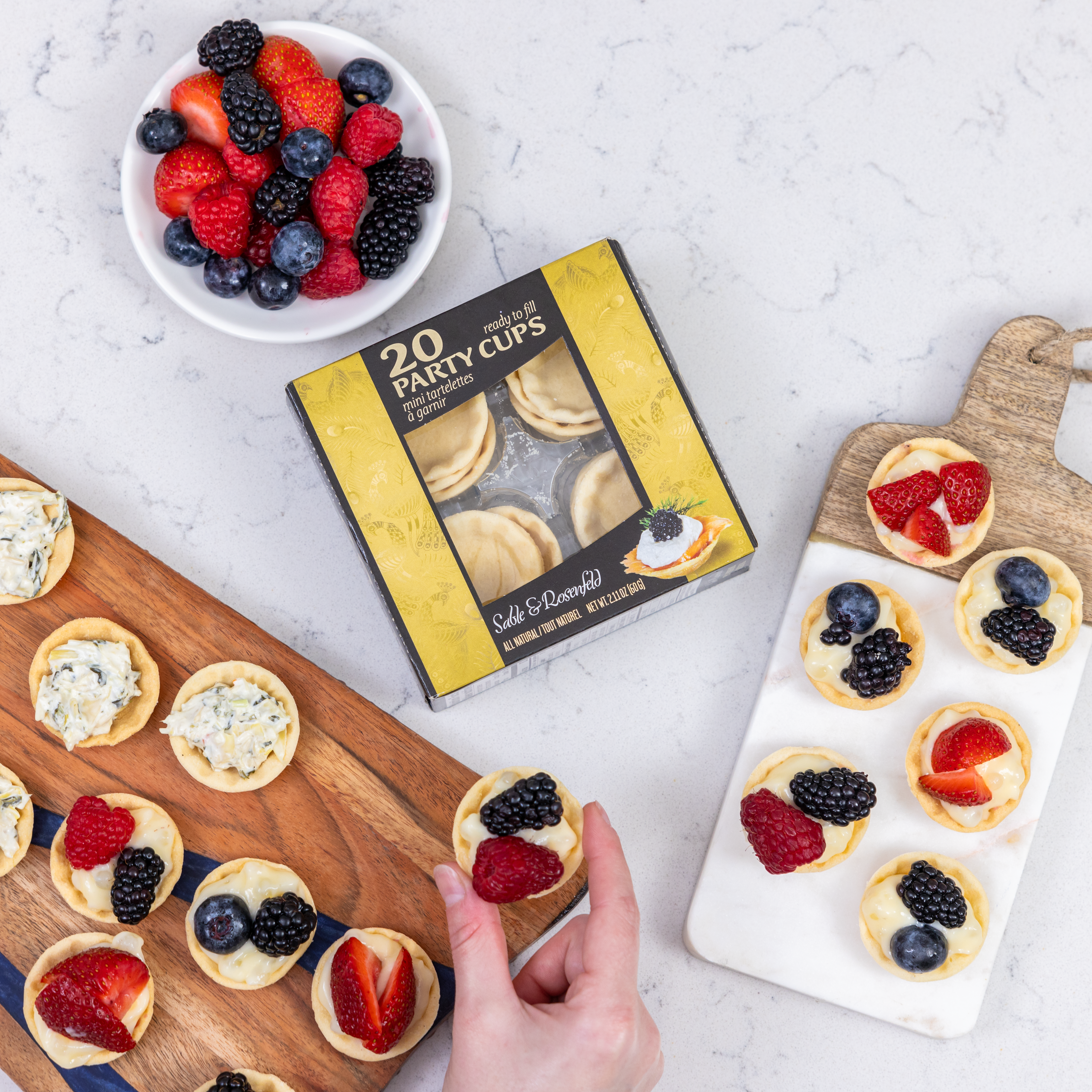 An assortment of mini tartelettes filled with cream and topped with fresh berries, alongside a box of Sable & Rosenfeld 20 Party Cups on a marble countertop.