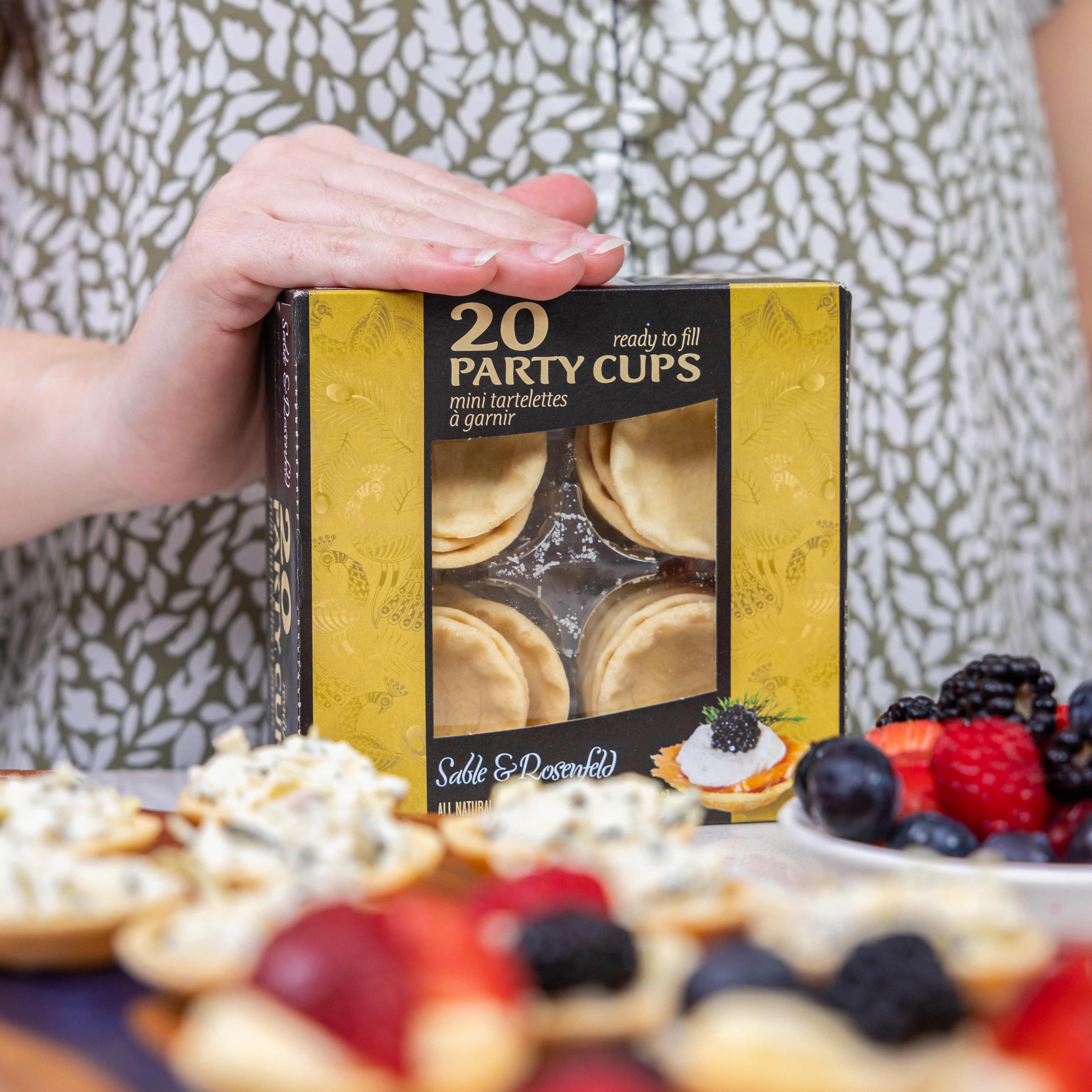 A person holding a box of Sable & Rosenfeld 20 Party Cups, surrounded by freshly prepared mini tartelettes filled with creamy toppings and fresh berries.