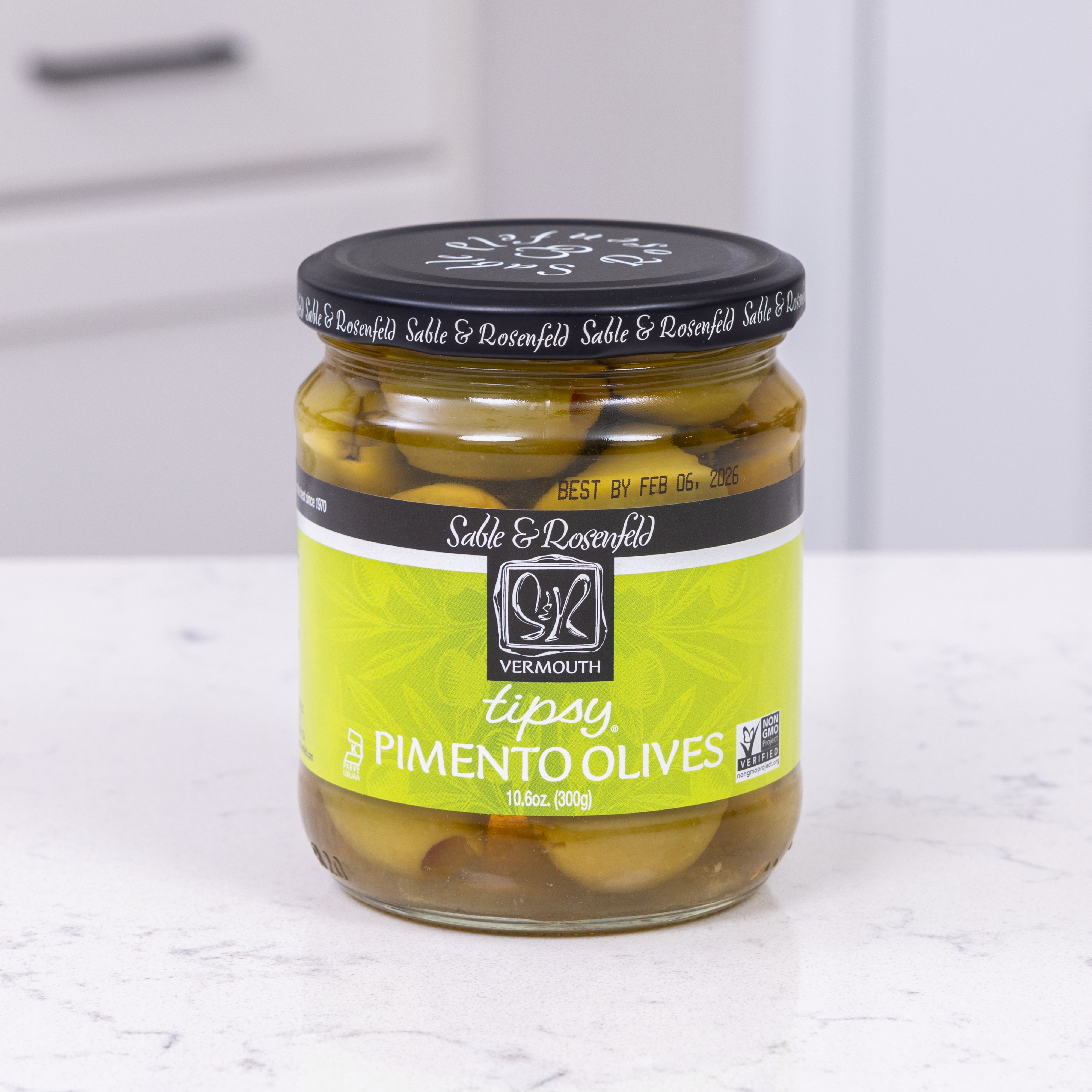 A jar of Sable & Rosenfeld Tipsy Pimento Olives infused with vermouth, featuring green olives stuffed with pimentos, placed on a white kitchen countertop.