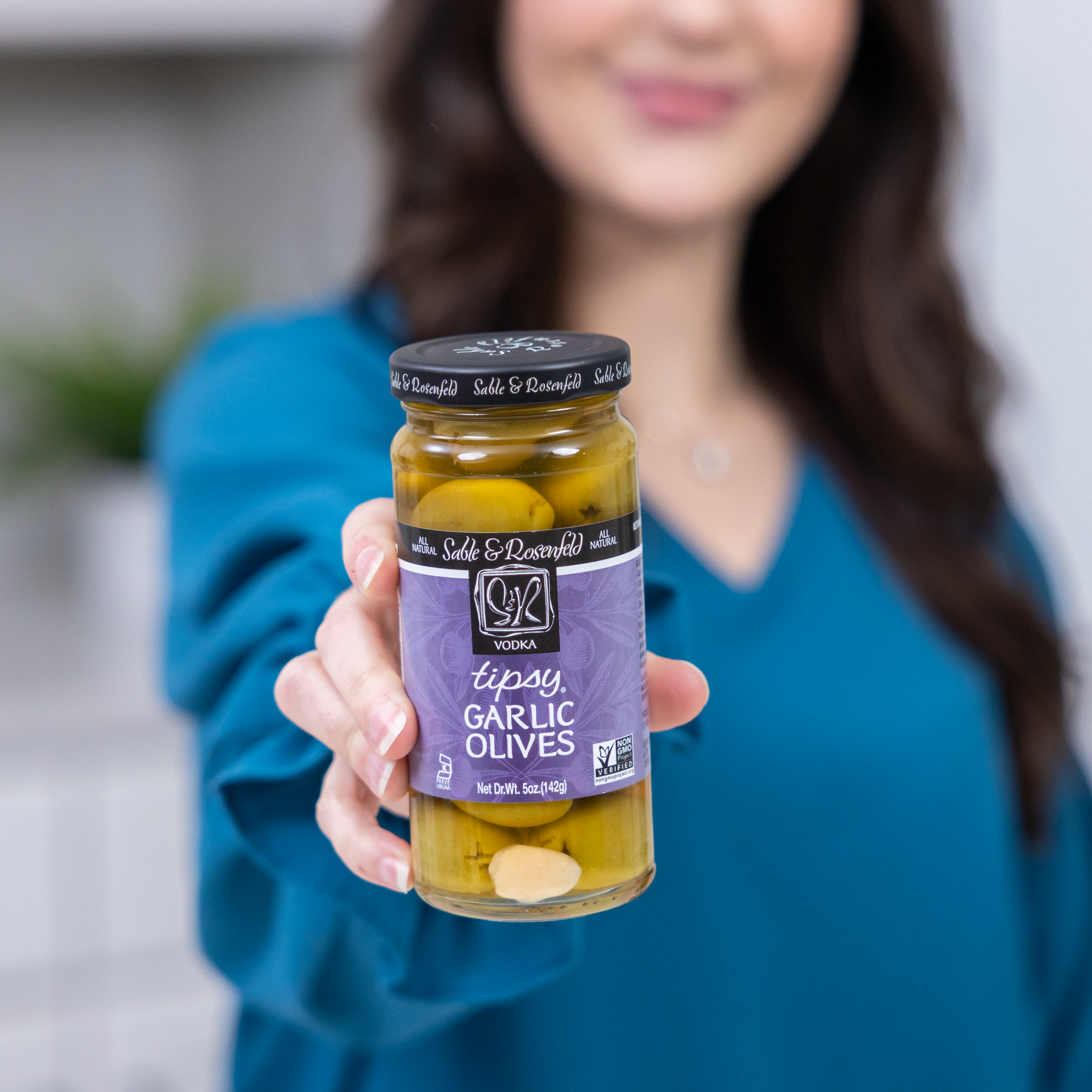 A woman in a blue blouse holding a jar of Sable & Rosenfeld Tipsy Fiery Olives, featuring green olives stuffed with garlic, in focus against a blurred background.