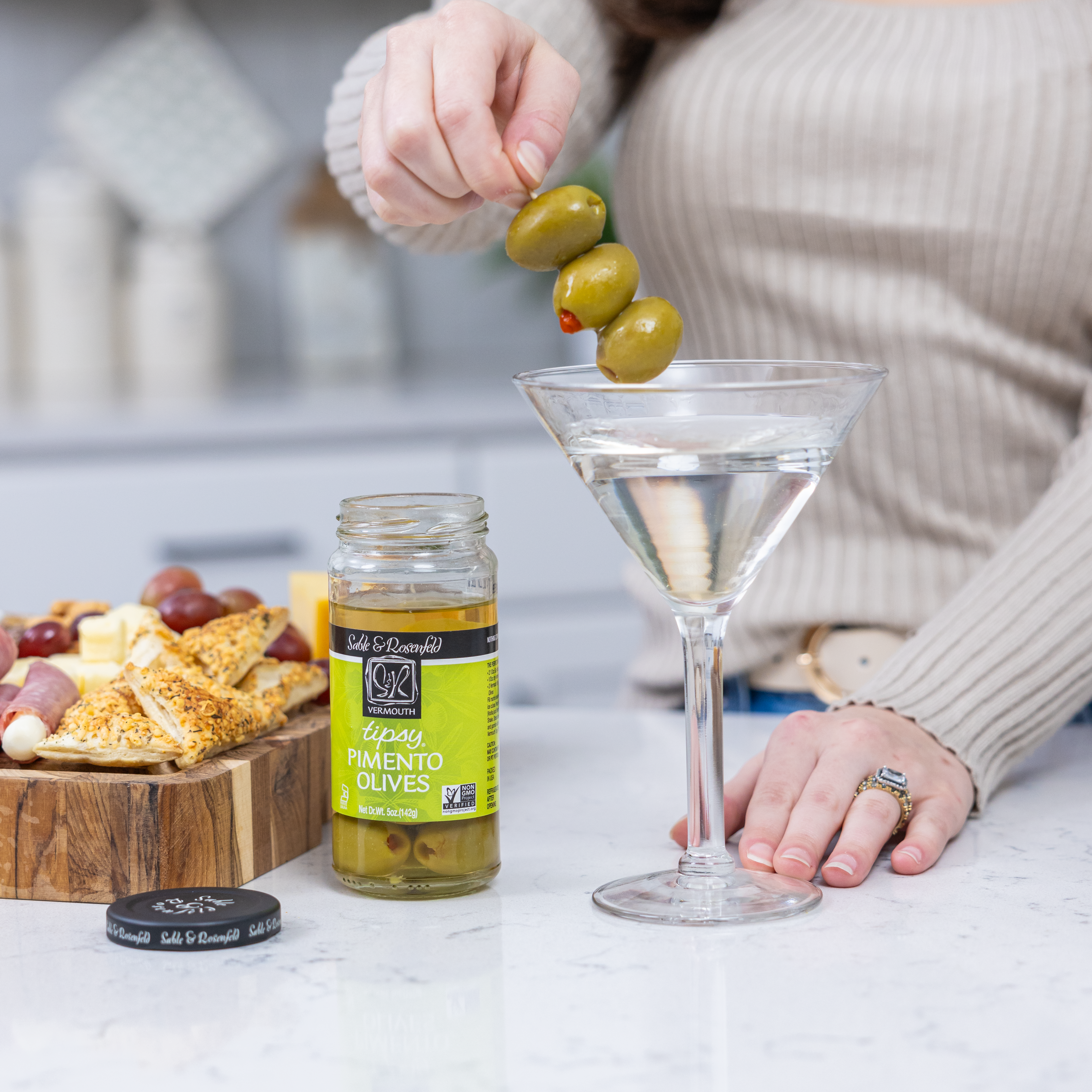 A jar of Sable & Rosenfeld Tipsy Pimento Olives on a white kitchen countertop, with a person garnishing a martini with skewered green olives, perfect for cocktails and charcuterie boards.