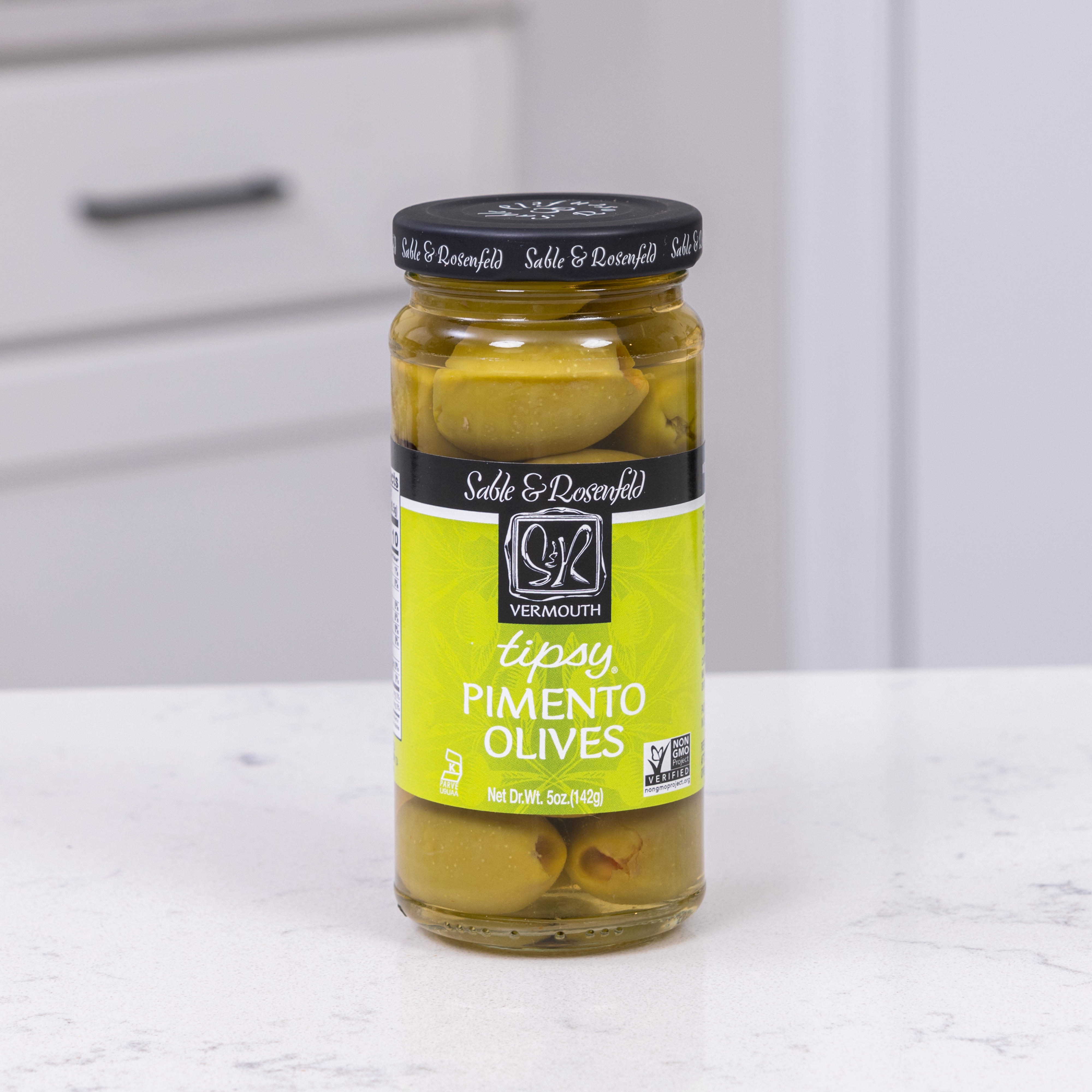 A jar of Sable & Rosenfeld Tipsy Pimento Olives infused with vermouth, featuring green olives stuffed with pimentos in brine, displayed on a white countertop with a modern kitchen background.