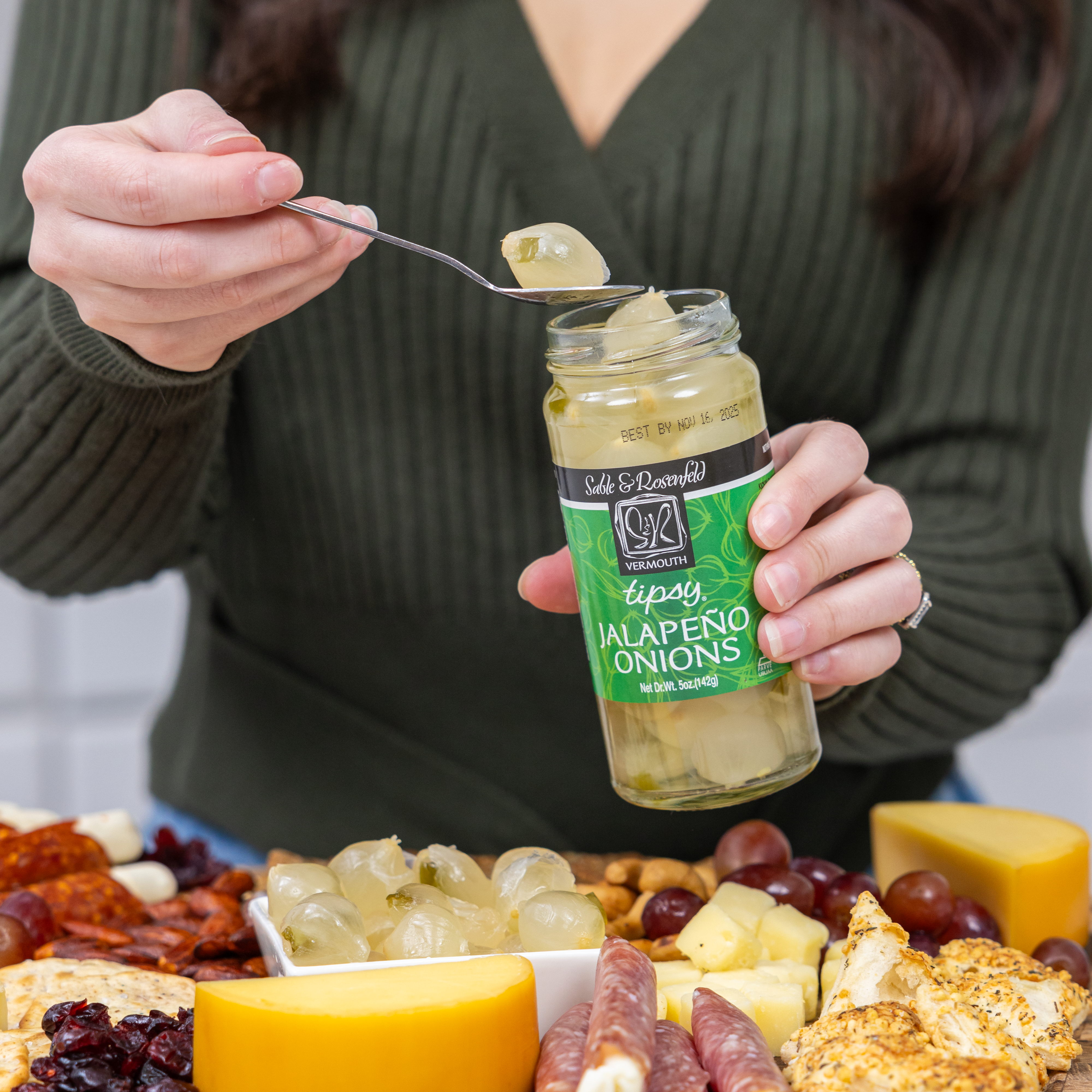 A person holding a jar of Sable & Rosenfeld Vermouth Onions while scooping out cocktail onions with a spoon over a charcuterie board filled with cheese, meats, nuts, and grapes.