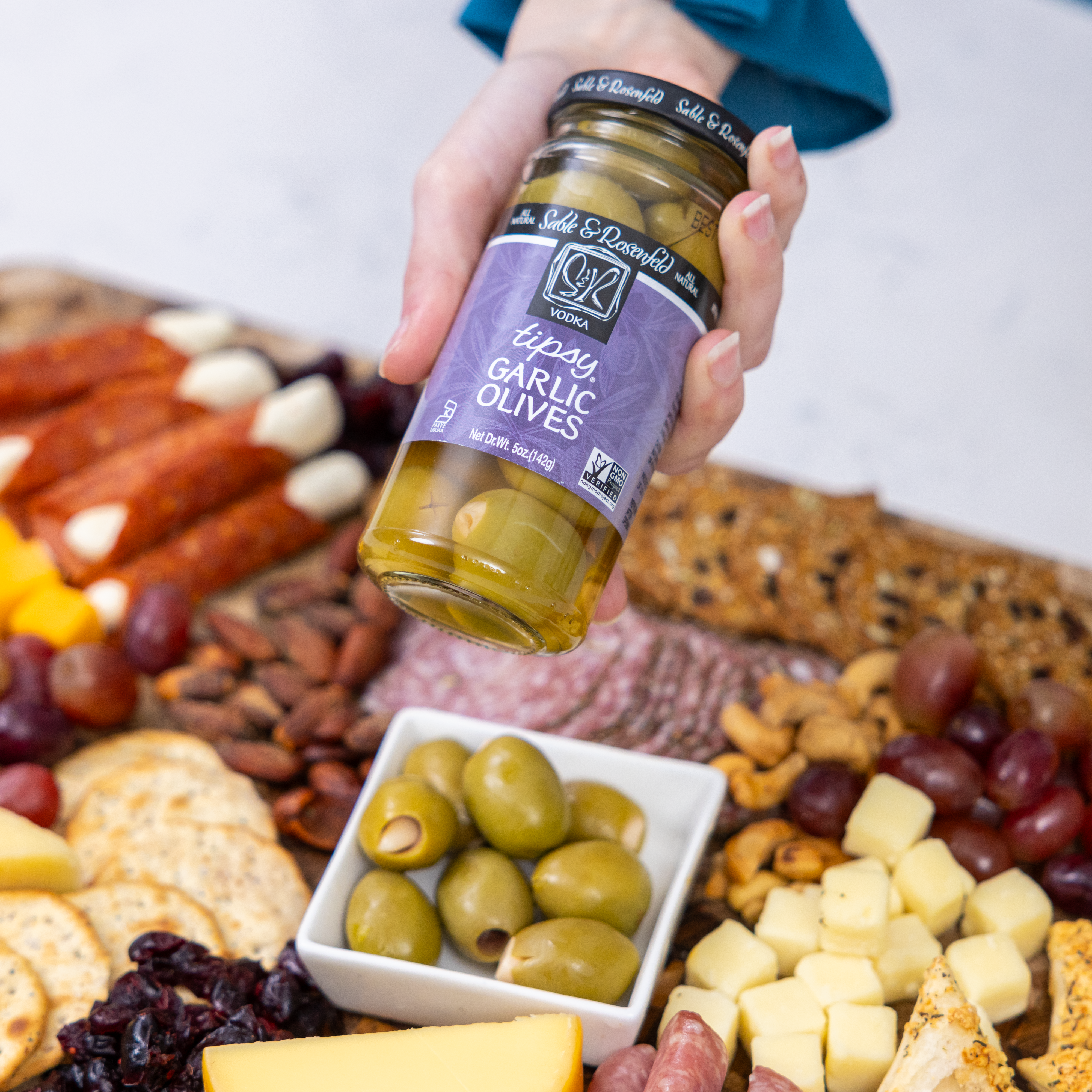 A person holding a jar of Sable & Rosenfeld Vodka Tipsy Fiery Olives above a charcuterie board featuring cheese, crackers, nuts, grapes, and a bowl of green olives.