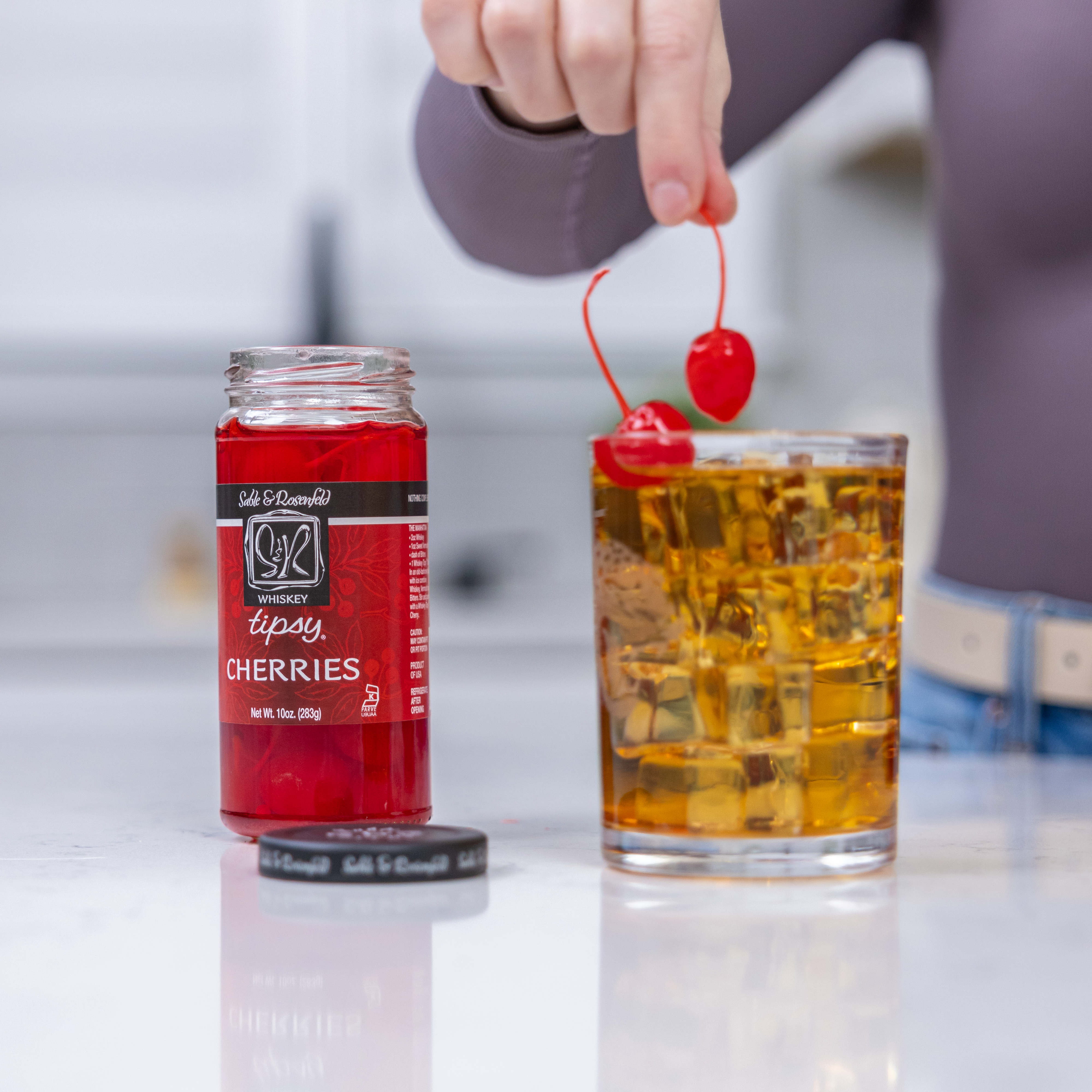 A hand garnishing a whiskey cocktail with vibrant red whiskey-infused cherries from a jar of Sable & Rosenfeld Whiskey Tipsy Cherries. The drink sits on a sleek white countertop, highlighting the rich amber tones of the whiskey and the gourmet appeal of the cherries.